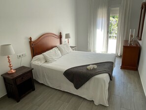 A neatly made bed with white linens and a dark throw blanket, a wooden nightstand with a lamp, and a window with curtains. - Budha's apartment, relax & confort center Calafell (Calafell)