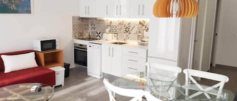 A compact living space with a kitchenette, a dining area with a glass table and white chairs, a red sofa, and a painting on the wall.