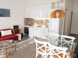 A compact living space with a kitchenette, a dining area with a glass table and white chairs, a red sofa, and a painting on the wall. - Budha's apartment, relax & confort center Calafell (Calafell)