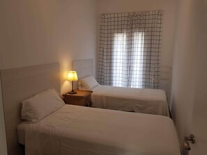 A hotel room with two beds, a nightstand with a lamp, and a window with a grid-patterned curtain. - Budha's apartment, relax & confort center Calafell (Calafell)