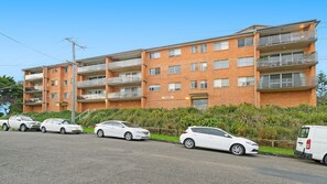 Un immeuble résidentiel à plusieurs étages, doté d’une façade en briques, de nombreux balcons et d’un espace de stationnement où sont garées plusieurs voitures.