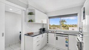 Une cuisine moderne avec des meubles blancs, un plan de travail noir et une fenêtre donnant sur des palmiers et des immeubles.