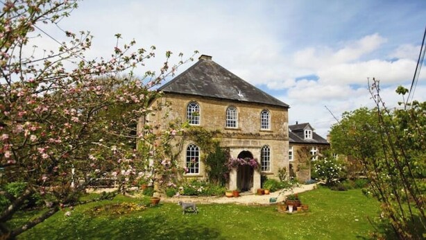 A stone building with a gabled roof, a central entrance with a door, and a small garden area with grass and flowering plants. - Chapel Cottage, spacious light and airy cottage in the Cotswolds AONB (Cirencester)