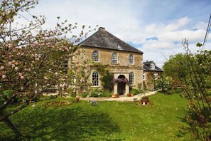 A stone building with a gabled roof, a central entrance with a door, and a small garden area with grass and flowering plants. - Chapel Cottage, spacious light and airy cottage in the Cotswolds AONB (Cirencester)