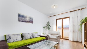Een moderne woonkamer met een glazen salontafel, een bruine bank met groene kussens, een witte fauteuil en een groot raam met gordijnen.