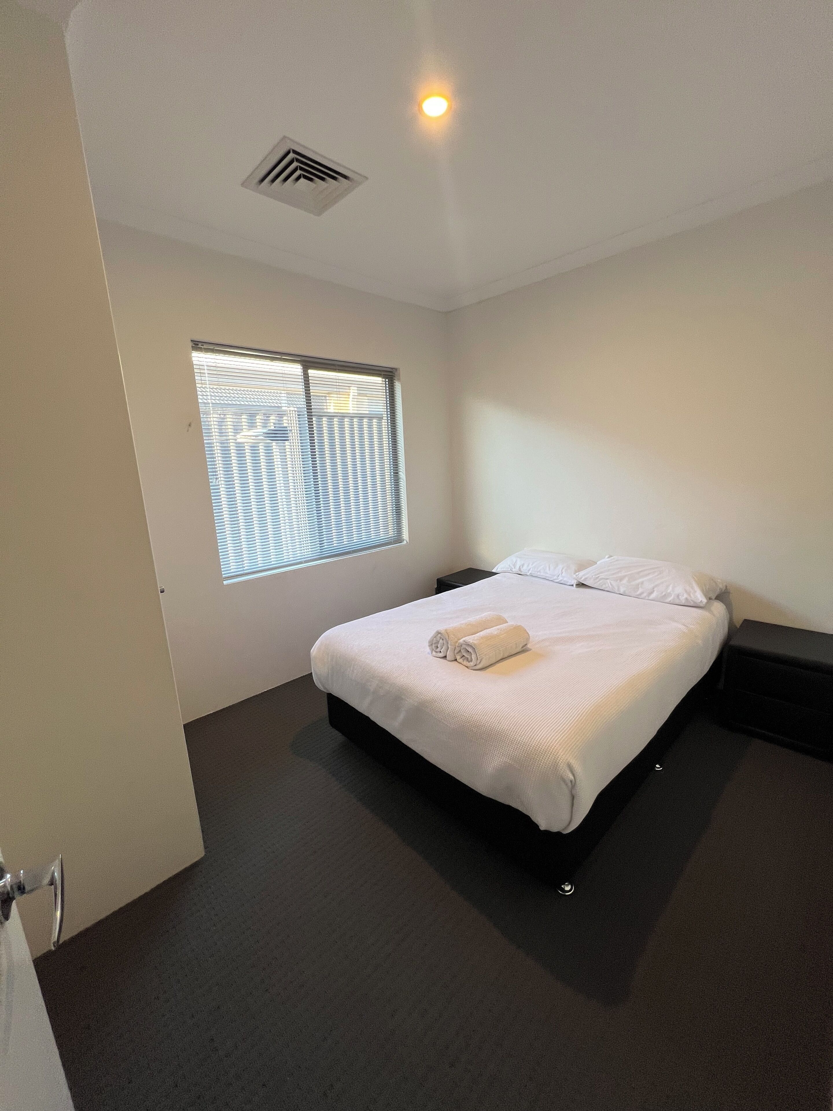 A hotel room with a bed, a window with blinds, a ceiling vent, and a nightstand.