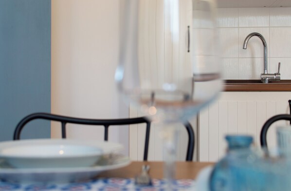 Una mesa de comedor con un plato y una copa, un jarrón azul, y una cocina al fondo con un lavabo y armarios.