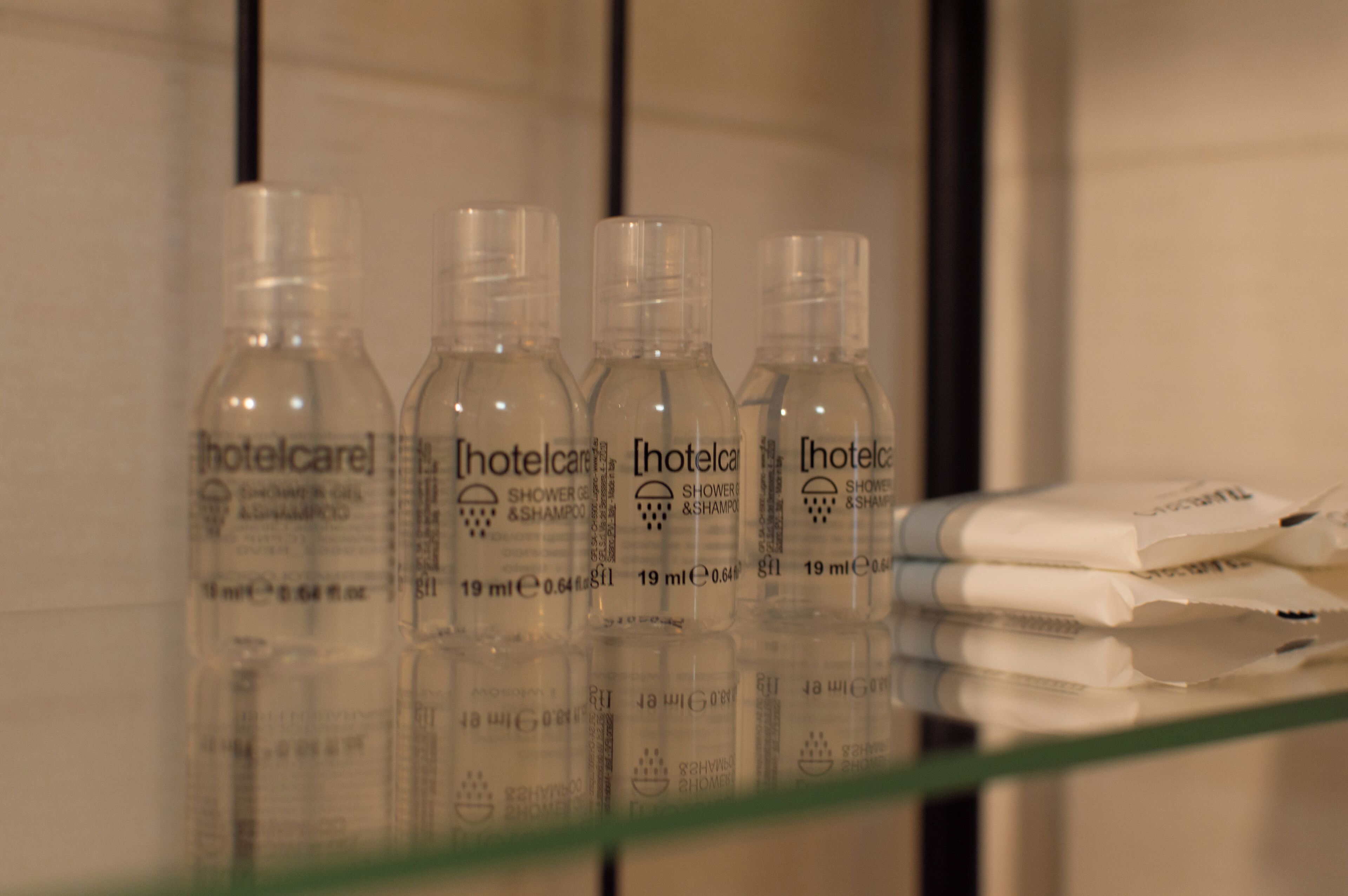 Quatre bouteilles de gel douche et de shampoing de la marque Hotelcare sont posées sur une étagère en verre.