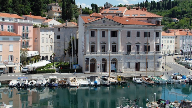 A marina with boats docked, a large building with classical architecture, and a hilly background with greenery.
