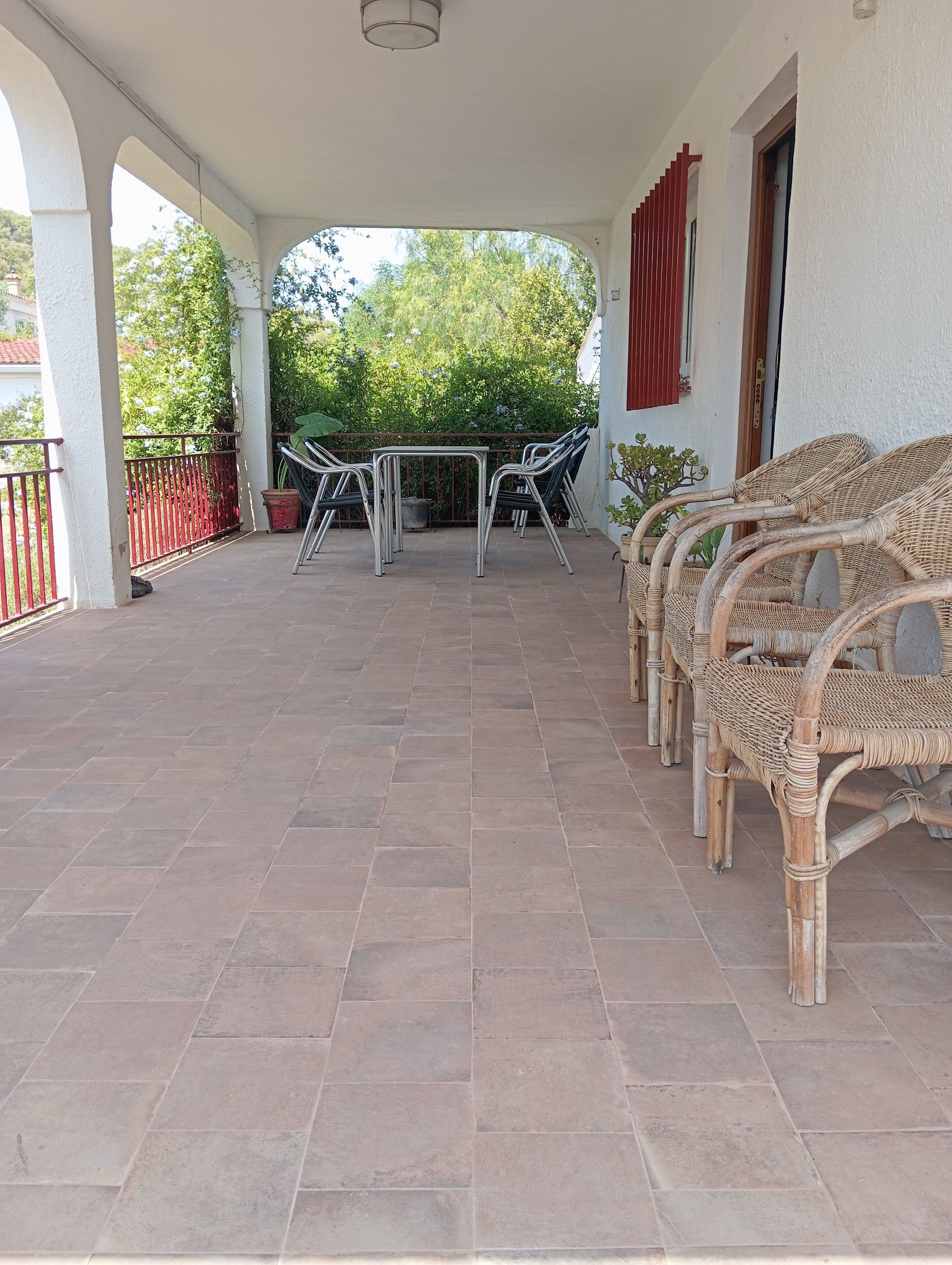 A covered patio with tiled flooring, metal-framed tables, wicker chairs, and a red railing.