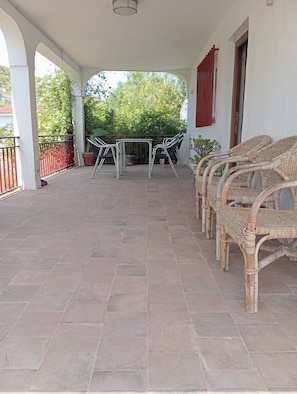 Un patio cubierto con piso de baldosas, mesas de estructura metálica, sillas de mimbre y barandal rojo.