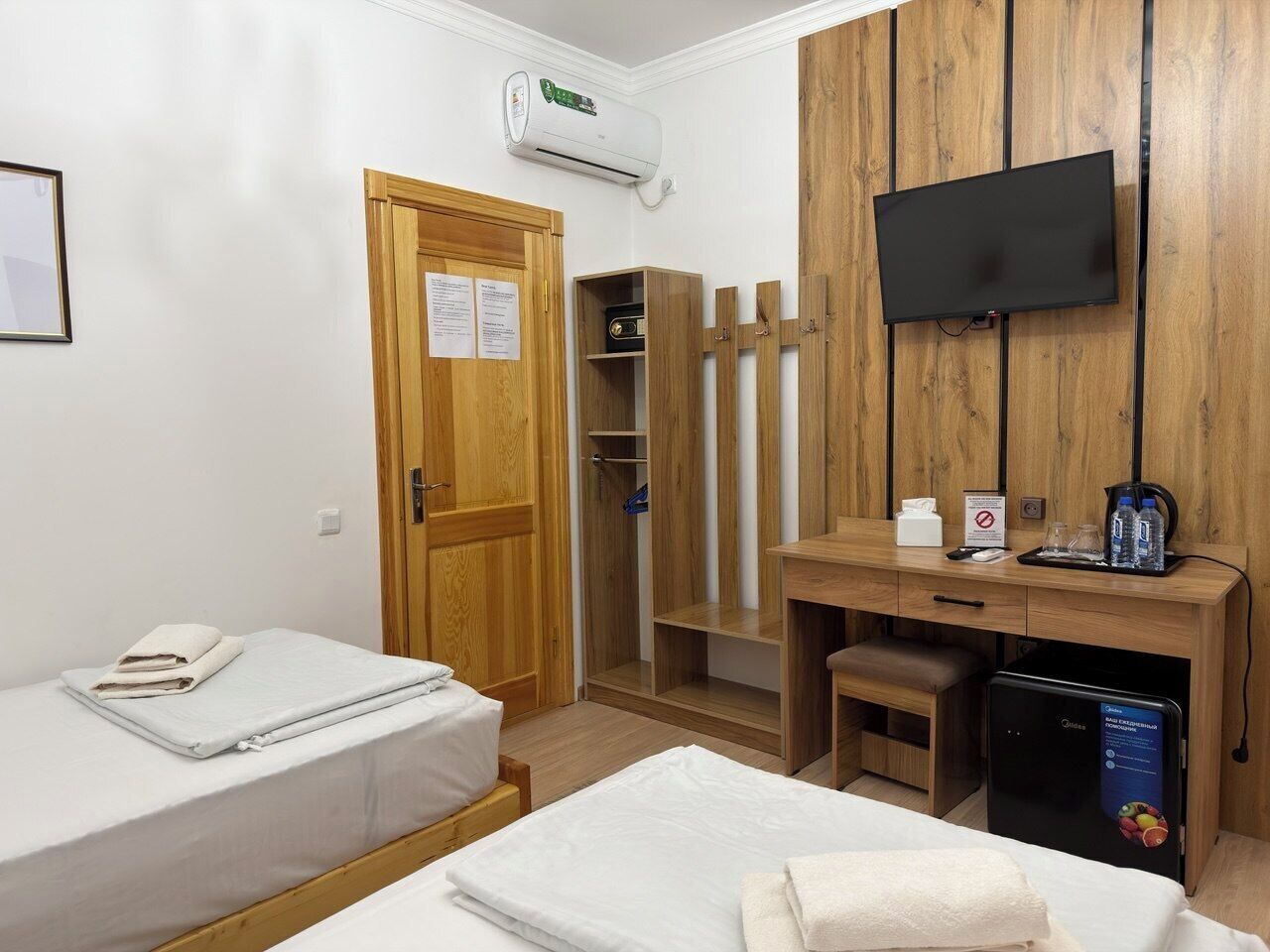 A hotel room with two beds, a wooden wardrobe, a desk with a computer, a TV, and an air conditioning unit.