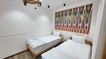 Una habitación pequeña y limpia con dos camas individuales, un cabecero de madera, un cuadro abstracto y colorido en la pared y una lámpara en el techo.