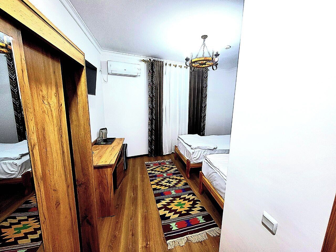 A narrow hotel room with two beds, a wooden desk, a patterned rug, and a chandelier.
