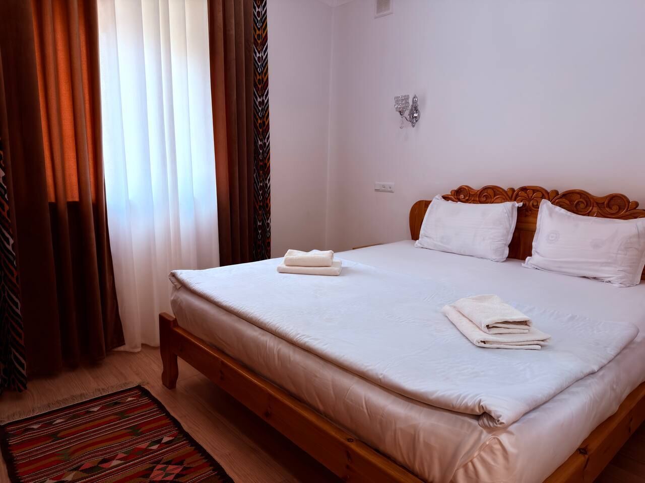 A bedroom with a wooden bed, white linens, a patterned rug, and curtains.