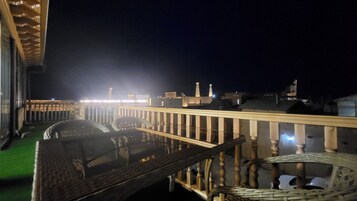 Vista nocturna de un balcón con mobiliario de mimbre y barandilla.