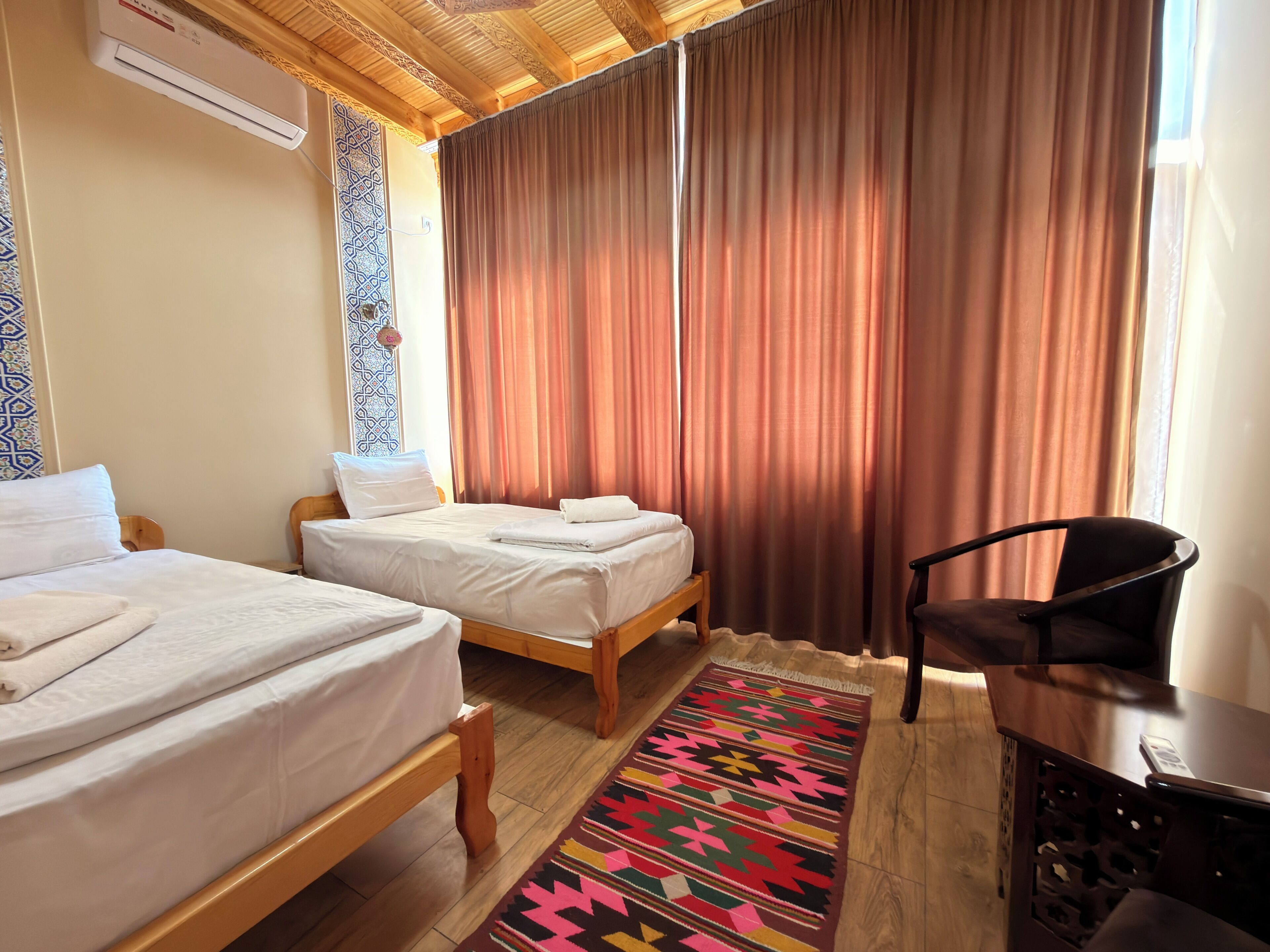 A hotel room with two beds, a desk, a chair, a patterned rug, and a window with curtains.