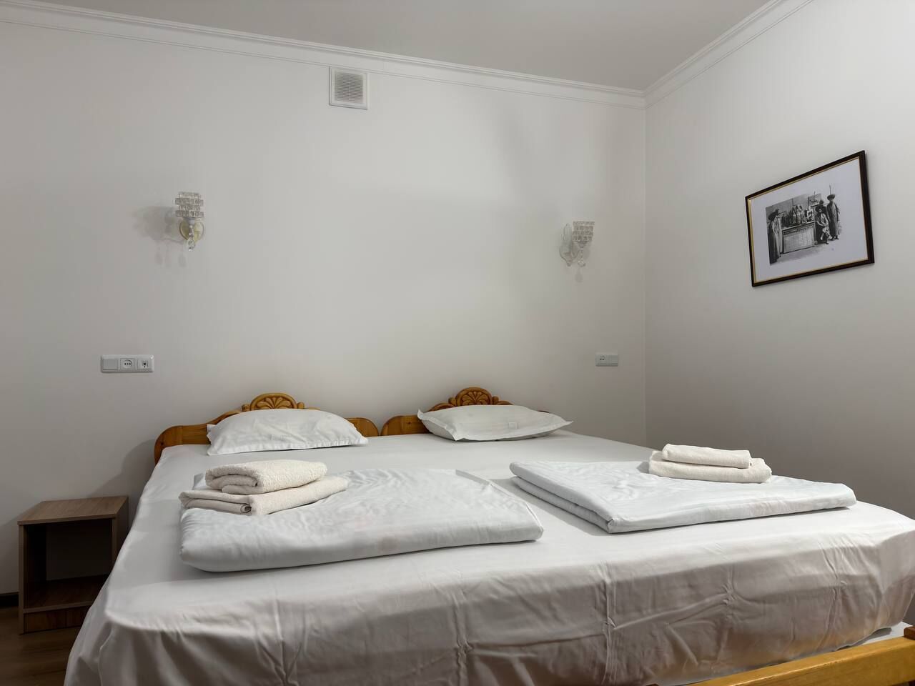 Un lit double bien fait, avec du linge de lit blanc et des serviettes pliées, une petite table de chevet et un tableau encadré au mur.