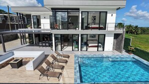 Una casa moderna de dos pisos con un amplio balcón de vidrio, una piscina y mobiliario de exterior.