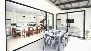 Una cocina moderna con armarios blancos, un comedor con mesa de vidrio y una escalera que lleva a un balcón.