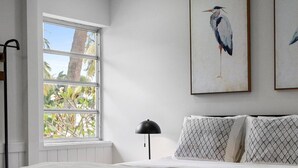A bedroom with a large window, a bed with patterned pillows, and a framed bird painting on the wall.