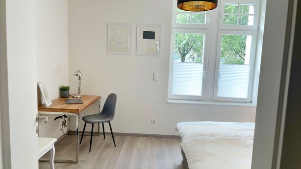A small, bright room with a wooden desk, a chair, a bed, and a window with blinds. - Apartment in guter Lage zur Innenstadt (Dortmund)