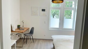 A small, bright room with a wooden desk, a chair, a bed, and a window with blinds. - Apartment in guter Lage zur Innenstadt (Dortmund)