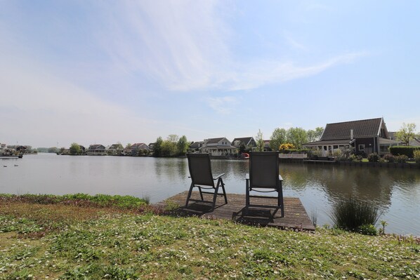 A lakeside view with two chairs on a dock, a row of houses, and a clear sky.