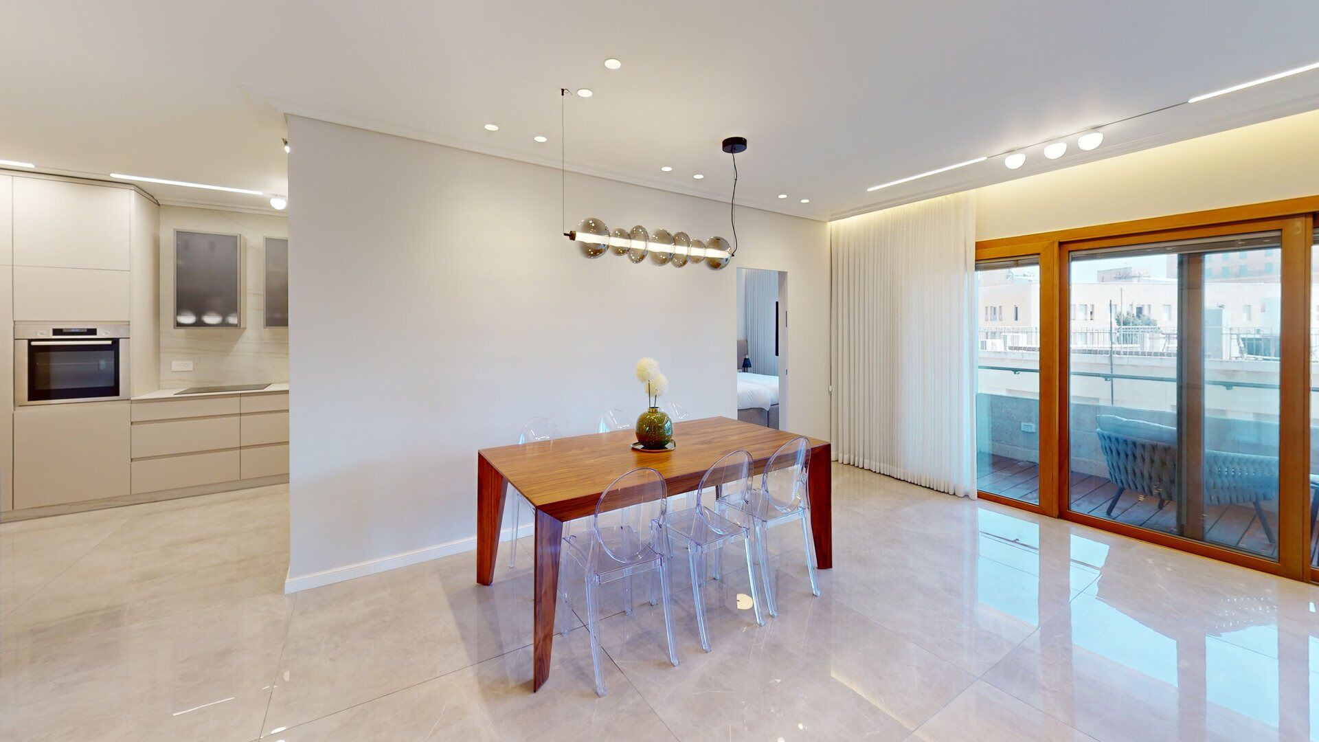 A modern kitchen with built-in appliances, a dining area with a wooden table and clear chairs, and a sliding glass door leading to a balcony.