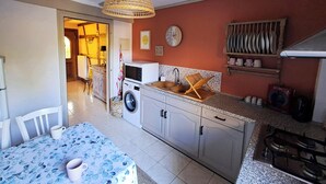 Een kleine keuken met een wasmachine, een spoelbak en een kookplaat. Er staat een eettafel met stoelen bij, en erboven hangt een lamp.