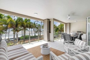 Ein modernes Wohnzimmer mit großem Fenster mit Blick auf einen Strand, einem Essbereich mit Sitzgelegenheiten im Freien und einer Küchenzeile.
