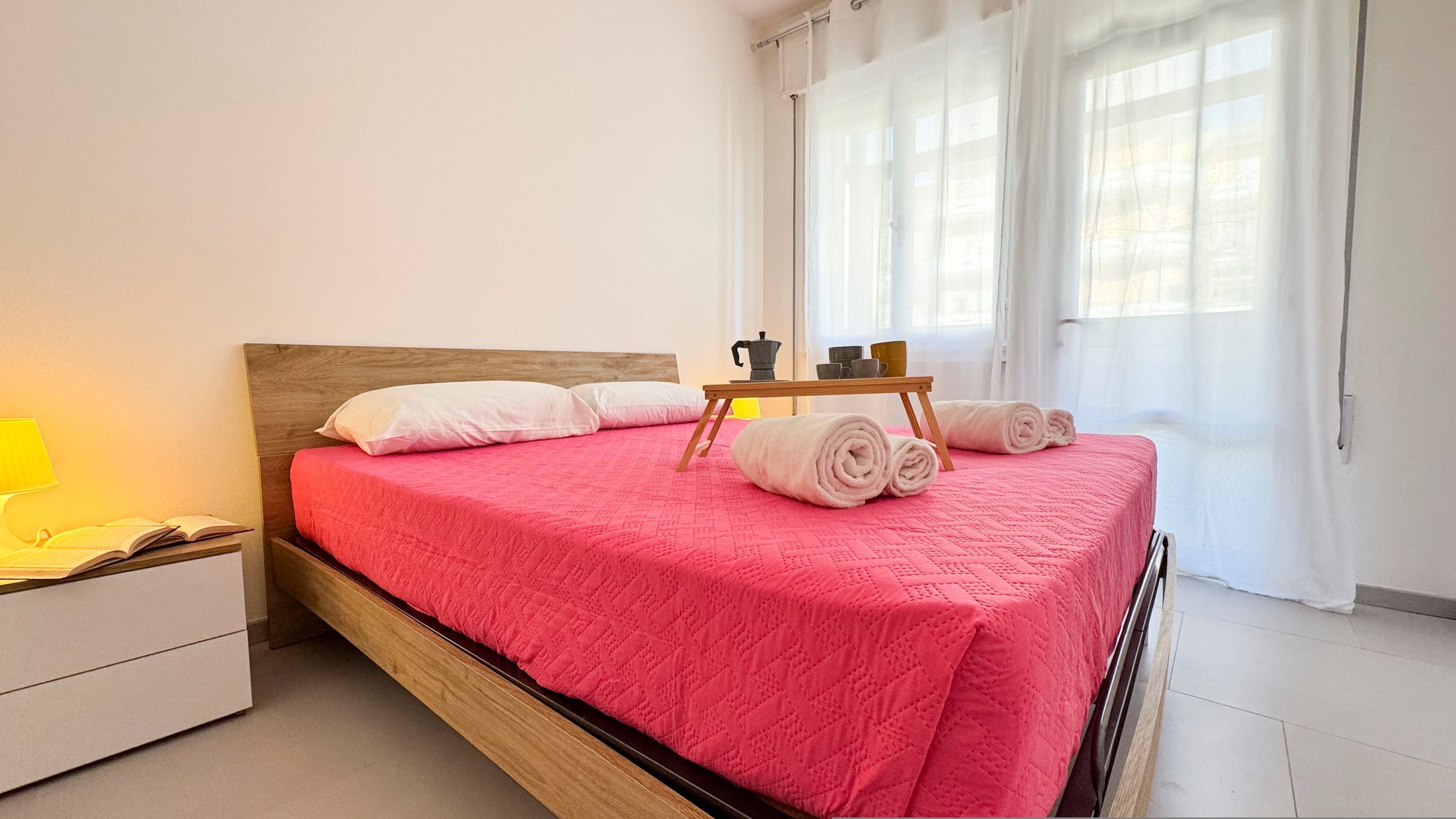 A neatly made bed with a pink quilt, a wooden headboard, and a bedside table with a lamp and an open book.