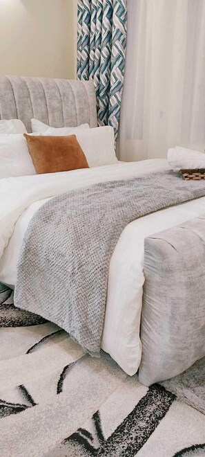 Una cama bien hecha con un plaid gris de punto, almohadas blancas y una almohada decorativa marrón.