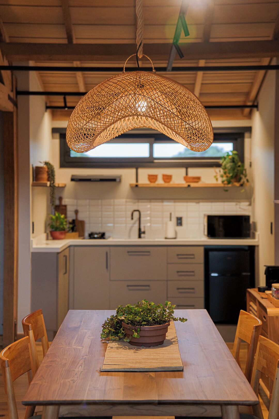 Et moderne køkken med et træspisebord, et hængt vævet lysarmatur og en potteplante på bordet.