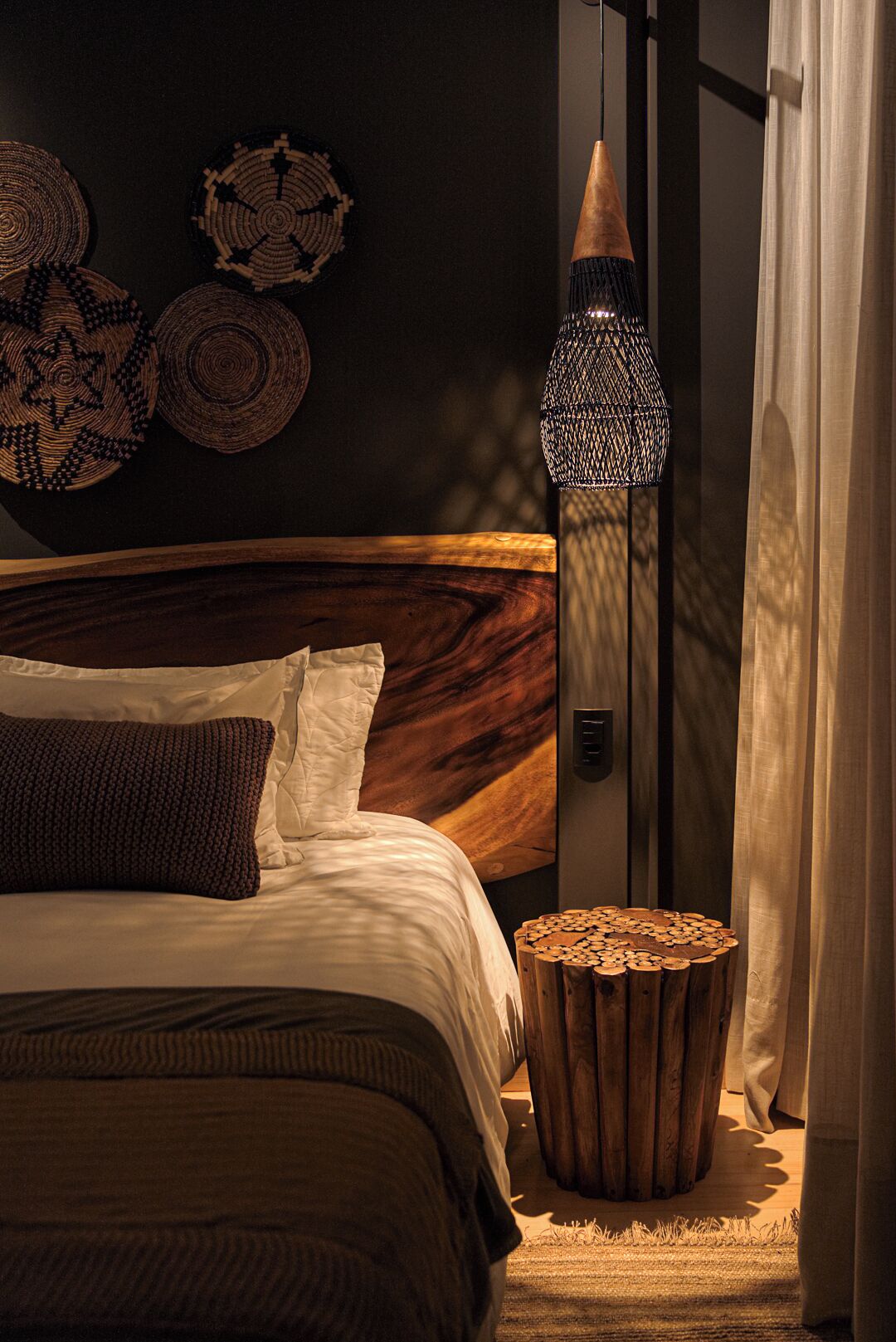 Un dormitorio con cabecera de madera, cama con ropa de cama blanca y marrón, lámpara de pie tejida y un taburete de madera pequeño.
