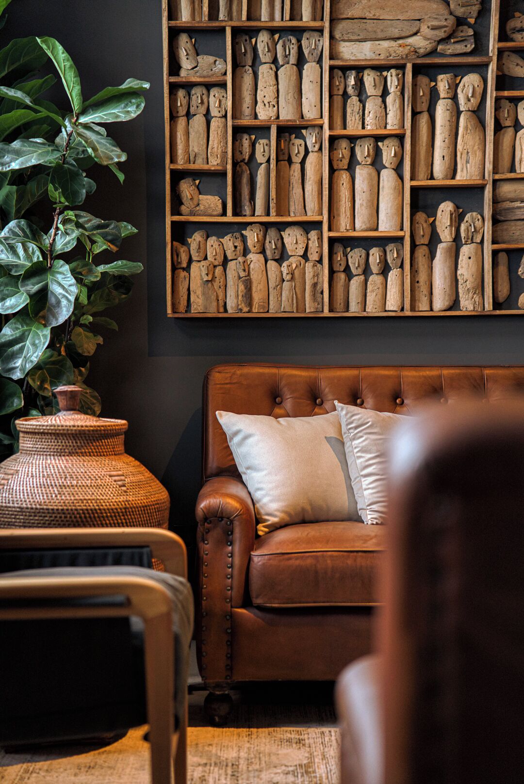 A room with a brown leather sofa, a large wooden sculpture wall, and a large green plant.