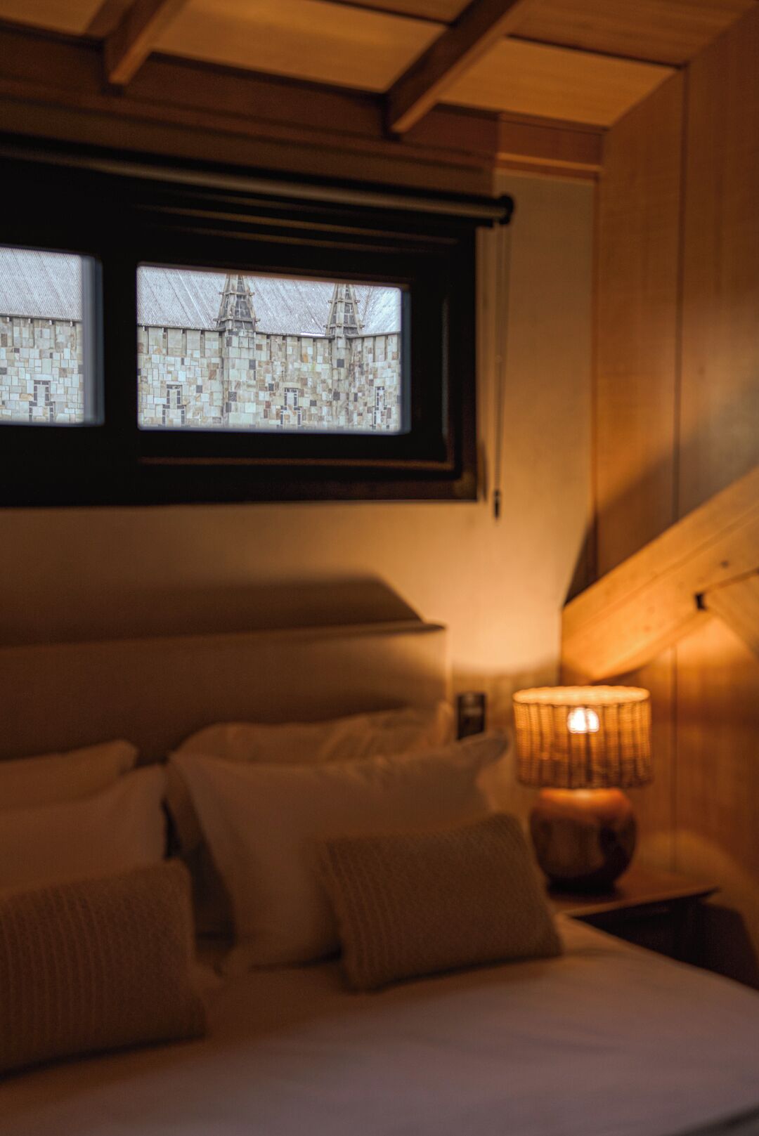 A bedroom with a bed, pillows, a nightstand, a lamp, and a window with a view of a building.