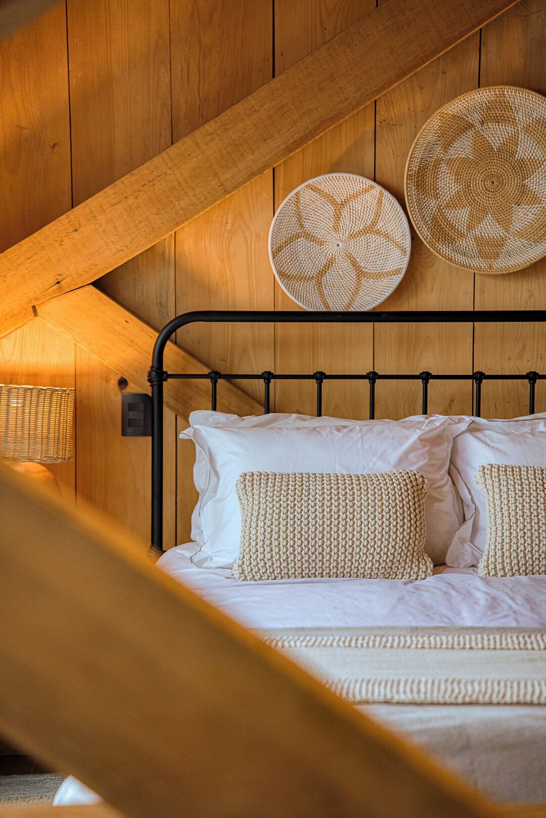 Techo de madera con diseño inclinado, cama con sábanas blancas y almohadas tejidas, y dos tapices circulares tejidos.