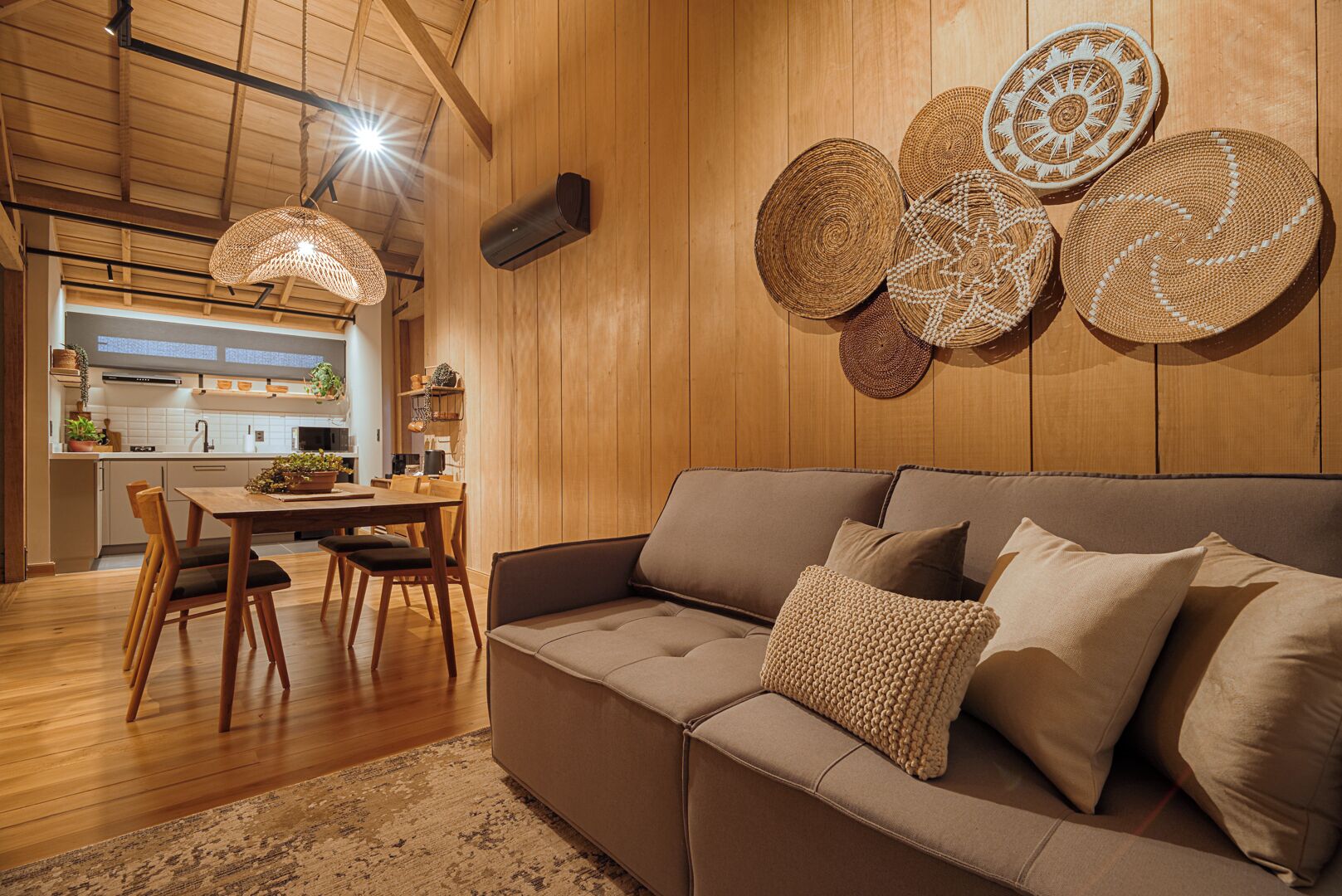 A cozy living room with a brown sofa, wooden dining table, and chairs. There are woven wall hangings and a hanging light fixture.