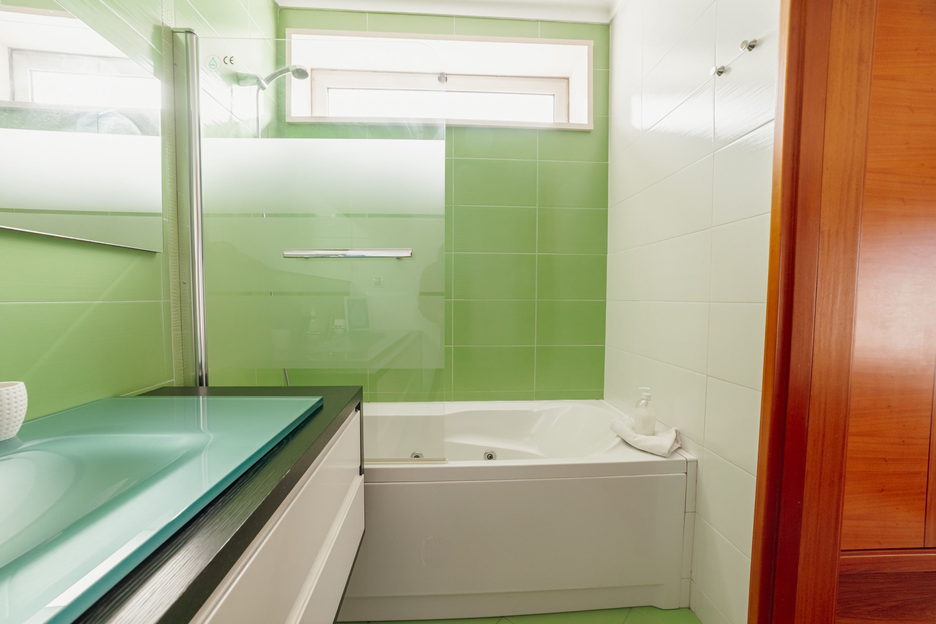 Ein Badezimmer mit Grün-Weiß-Farbgebung, einer Duschkabine aus Glas, einer weißen Badewanne und einer Holztür.
