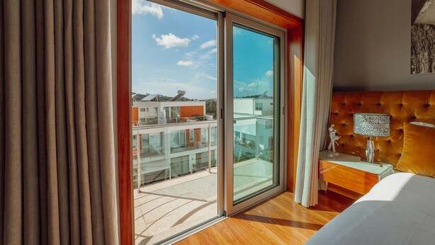 Una habitación de hotel moderna con un gran ventanal corredero a un balcón, un cabecero capitonado y una mesita de noche con lámpara.