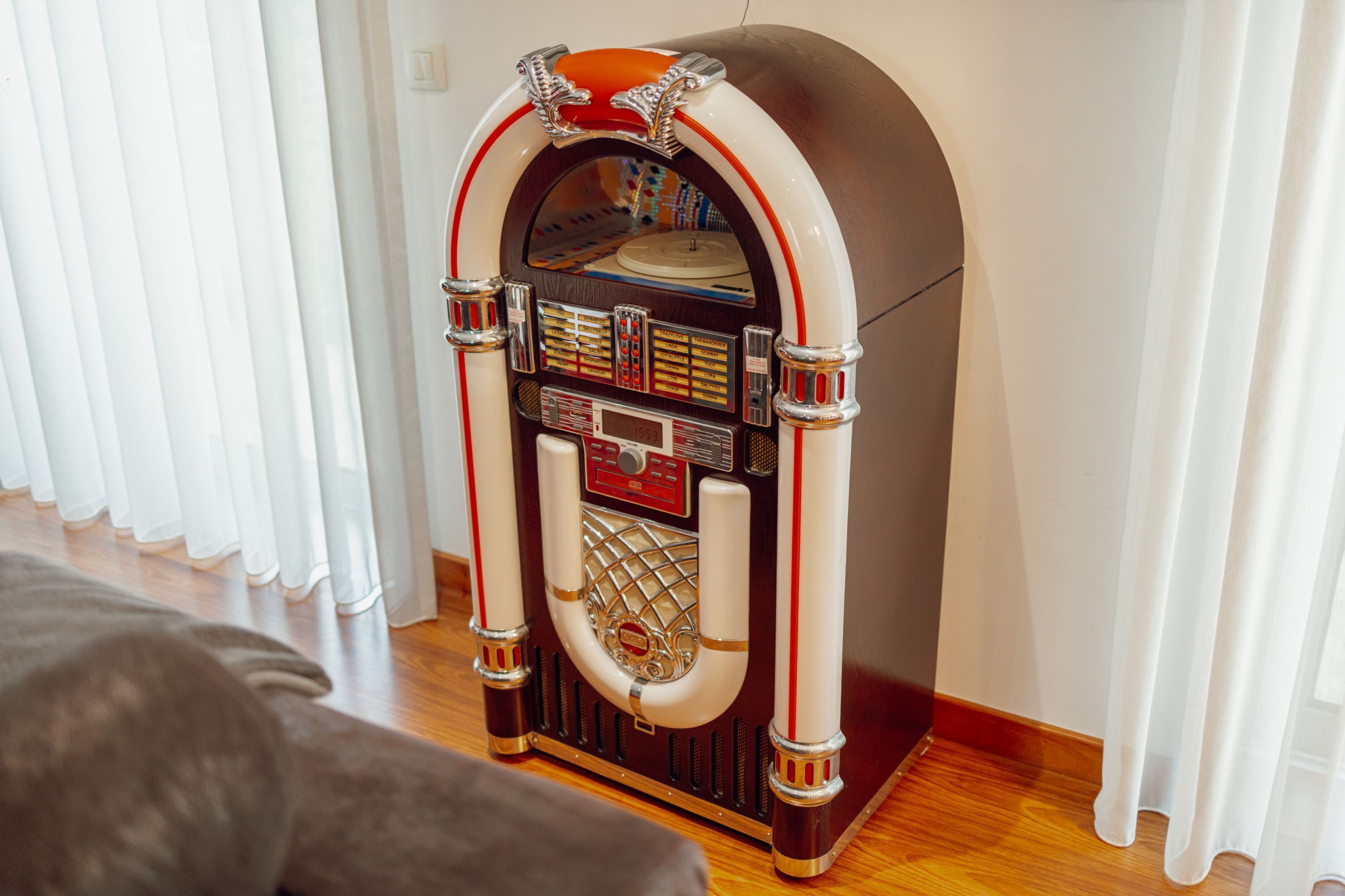 Een klassieke jukebox in een ruimte met houten vloeren en witte gordijnen.