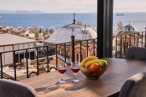 A table with a fruit bowl and two glasses of red wine, overlooking a coastal cityscape with domed buildings.