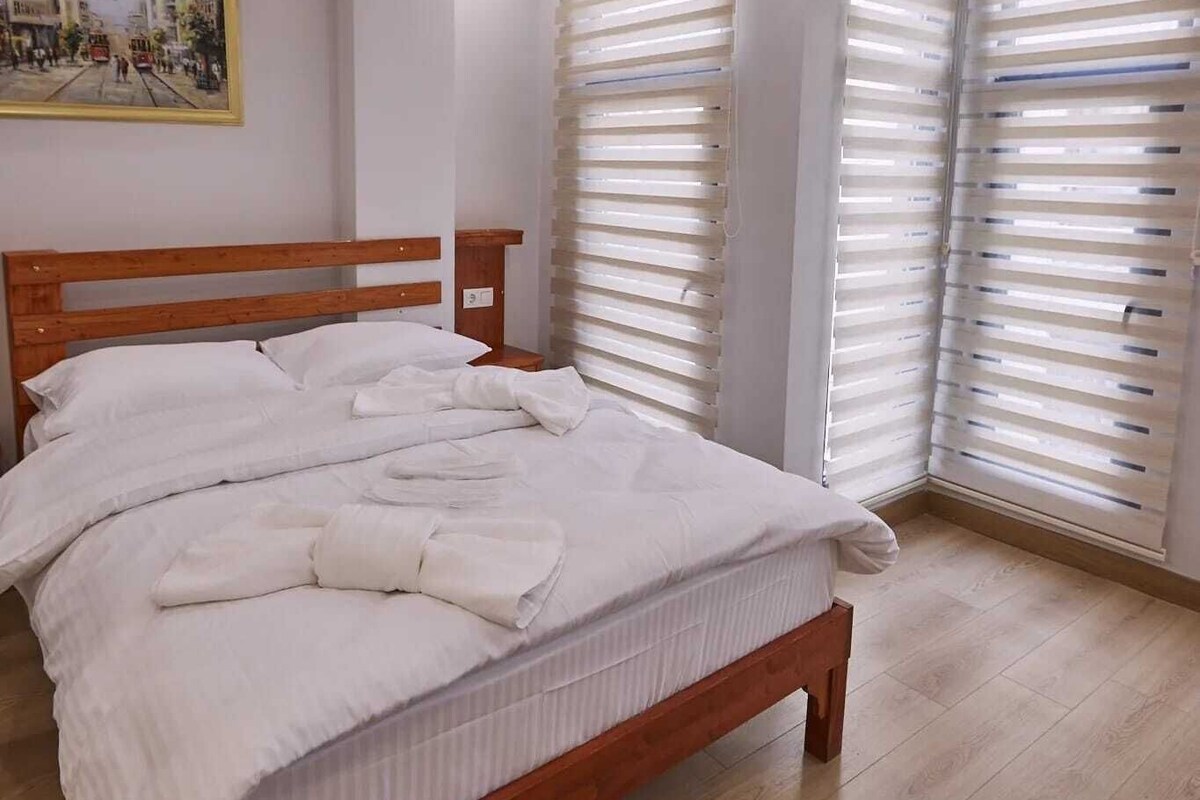 Une chambre à coucher avec un lit en bois, du linge de lit blanc, un tableau représentant un paysage urbain et une fenêtre avec des stores.