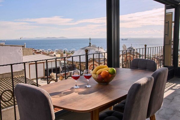 Une table à manger avec des chaises, un saladier de fruits et deux verres à vin sur un balcon donnant sur une vue côtière.