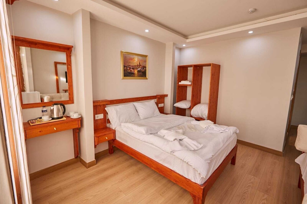 Un lit bien fait dans une chambre d’hôtel, avec une tête de lit en bois, une table de chevet sur laquelle se trouvent une bouilloire et des tasses, un miroir et un tableau accroché au mur.