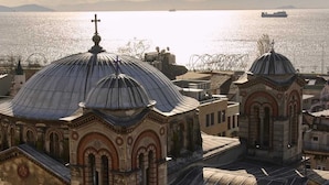 Vista de una iglesia con cúpulas y una cruz en la parte superior, con vistas a una masa de agua donde se distingue un barco a lo lejos.