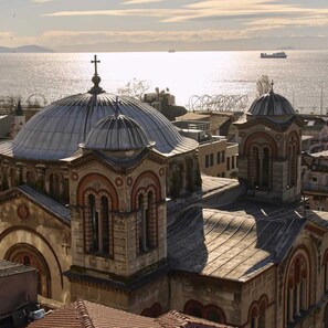 Une vue d’une église dotée de toits en forme de dôme et surmontée d’une croix, surplombant une étendue d’eau où l’on aperçoit un navire au loin.