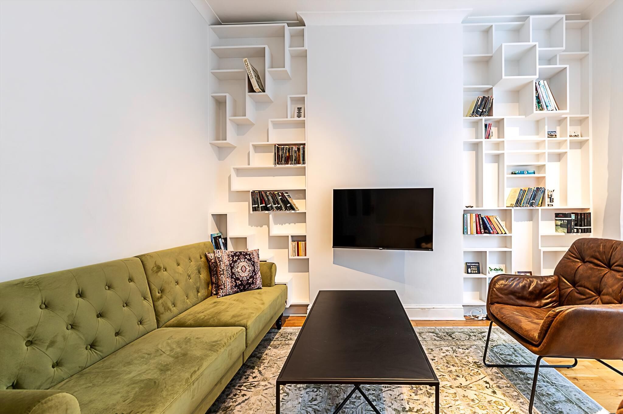 Uma sala de estar moderna com um sofá verde, uma poltrona de couro marrom, uma mesa de centro preta e estantes brancas com livros.