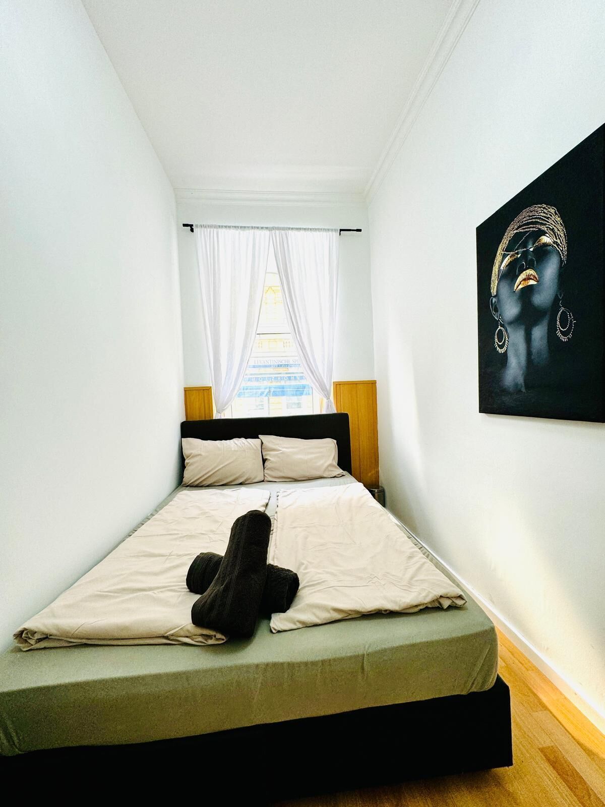 A small, modern bedroom with a single bed, a large window with sheer curtains, and a painting on the wall.